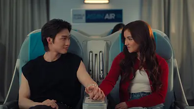 Kitty und Minho sitzen im Staffelfinale von „XO, Kitty“ in einem Flugzeug und halten Händchen. - Foto: Courtesy of Netflix