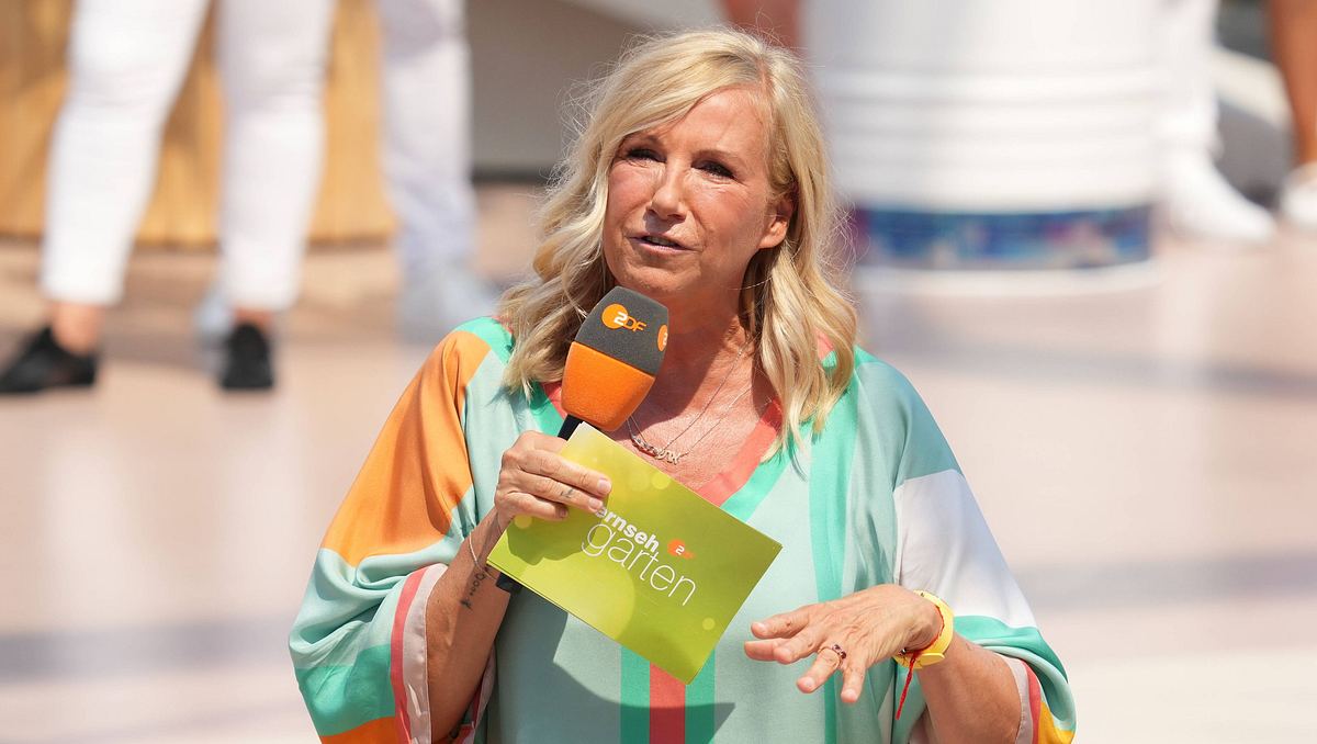 Andrea Kiewel steht in buntem Outfit, Karteikarte und Mikrofon auf der Bühne des ZDF-„Fernsehgarten“. Ihr Blick ist ernst. 