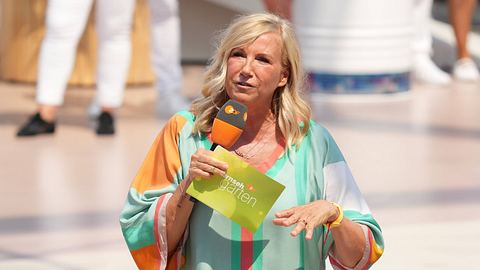 Andrea Kiewel steht in buntem Outfit, Karteikarte und Mikrofon auf der Bühne des ZDF-„Fernsehgarten“. Ihr Blick ist ernst.  - Foto:  IMAGO / BOBO