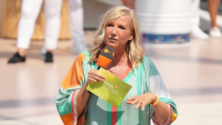 Andrea Kiewel steht in buntem Outfit, Karteikarte und Mikrofon auf der Bühne des ZDF-„Fernsehgarten“. Ihr Blick ist ernst.  - Foto:  IMAGO / BOBO
