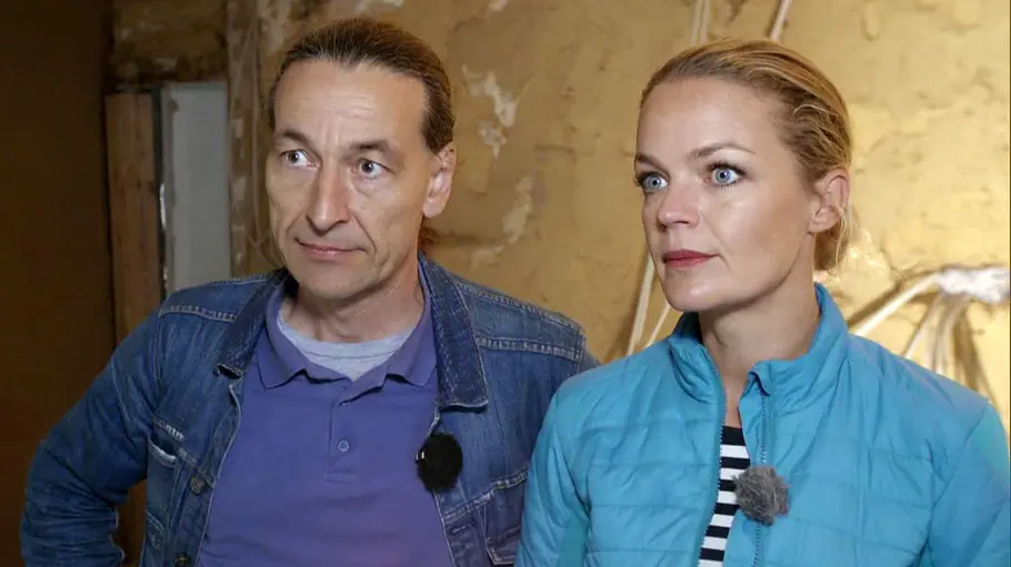 Zuhause im Glück-Absetzung Zuhause im Glück-John in Jeansjacke und Eva Brenner in türkisfarbener Steppjacke stehen mit Ansteckmikrofonen vor einer unverputzten Wand.