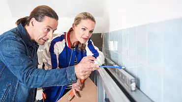 Szene aus Zuhause im Glück. John zeigt mit einem Bleistift auf eine hellblau geflieste Wand über einer Wasserwaage, während Eva daneben zusieht. - Foto: RTL II Szene aus Zuhause im Glück. John zeigt mit einem Bleistift auf eine hellblau geflieste Wand über einer Wasserwaage, während Eva daneben zusieht. - Foto: RTL II
