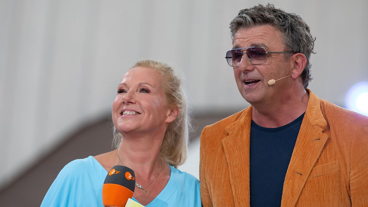 Zumindest Andrea Kiewel wirkte nicht so, als hätte sie ein Problem mit Hans Sigl. Andrea Kiewel und Hans Sigl stehen nebeneinander und moderieren gemeinsam den ZDF Fernsehgarten. Kiewel hält ein ZDF-Mikrofon in der rechten Hand.